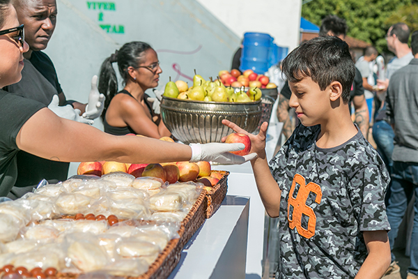 Funcionária entrega uma maçã a um garoto em um buffet de comida, com frutas e sanduíches em um parque.
