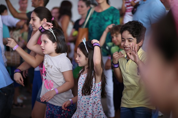 Diversas crianças felizes olham na mesma direção. Algumas mexem os braços e outras riem. Ao fundo, há vários adultos.