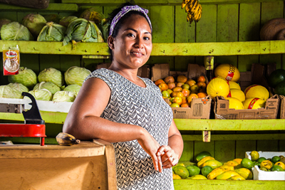Mulher com lenço nos cabelos posa para a foto em uma venda, ao lado de balcão, e há prateleiras com frutas ao fundo