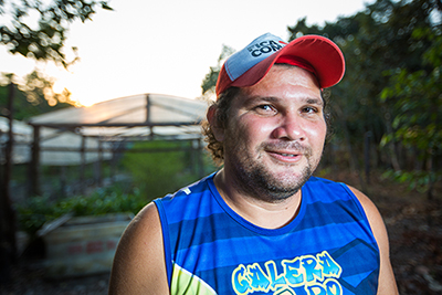 Homem de regata azul e boné posa para a foto sorrindo em um cenário de fazenda.