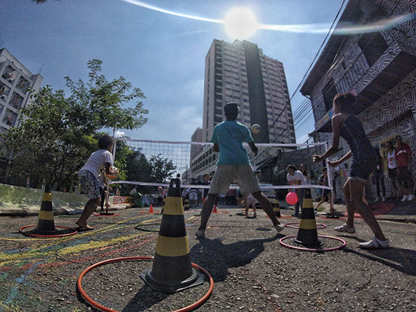 Crianças jogam vôlei ao ar livre sobre uma superfície de asfalto. Em volta há cones de trânsito com bambolês coloridos.