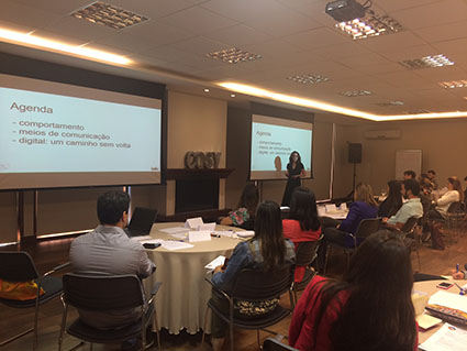 Uma mulher apresenta slides em um telão e grupos de pessoas assistem a redor de mesas redondas.