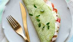 Prato com uma tapioca verde recheada de creme e tomate, com ervas frescas por cima, ao lado de um garfo e faca.