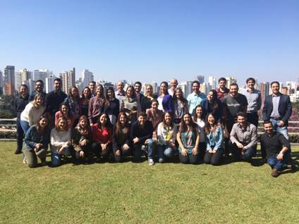 Grupo com cerca de 30 pessoas posa para foto em um gramado. Ao fundo, há um cenário urbano com prédios.