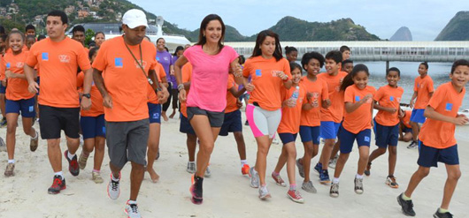 Adultos e crianças correm juntos na praia usando camisetas cor de laranja e bermudas azuis.