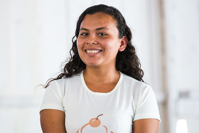 Mulher de cabelos castanhos ondulados está sentada sorrindo para a foto.