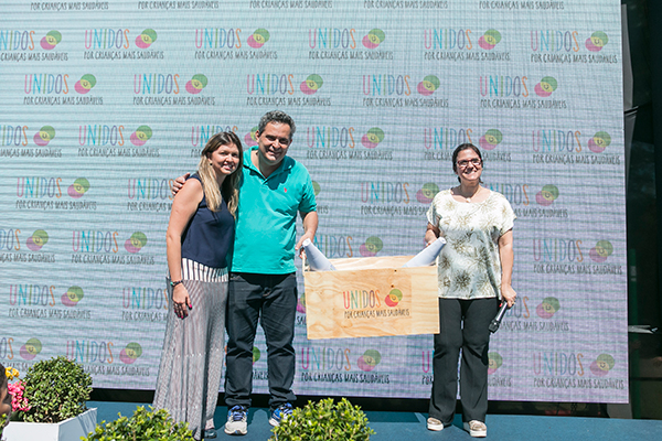 Um casal e uma mulher com microfone posam para foto com uma caixa de madeira com o logo Unidos por crianças mais saudáveis.