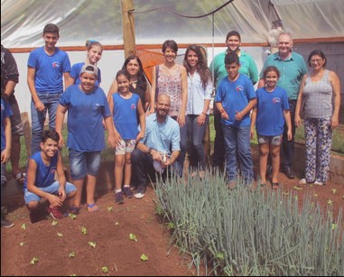 Crianças usando uniforme azul posam ao lado de alguns adultos em frente a uma horta